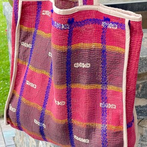 K&ouml;nnte beinhalten: Eine gro&szlig;e, gesteppte Tragetasche in Pink und Lila mit blauen und goldenen Verzierungen. Die Tasche hat ein kariertes Muster und ist aus einem weichen, strukturierten Stoff gefertigt.