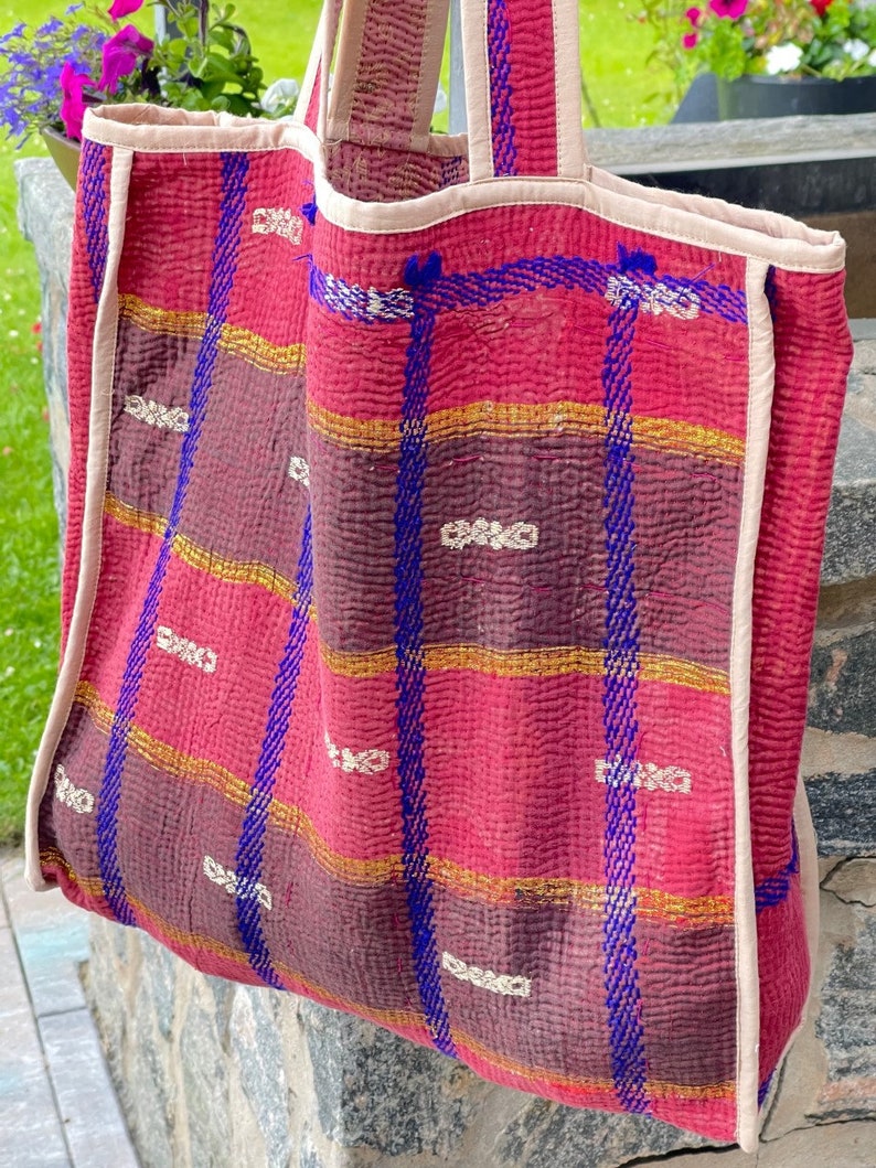 K&ouml;nnte beinhalten: Eine gro&szlig;e, gesteppte Tragetasche in Pink und Lila mit blauen und goldenen Verzierungen. Die Tasche hat ein kariertes Muster und ist aus einem weichen, strukturierten Stoff gefertigt.