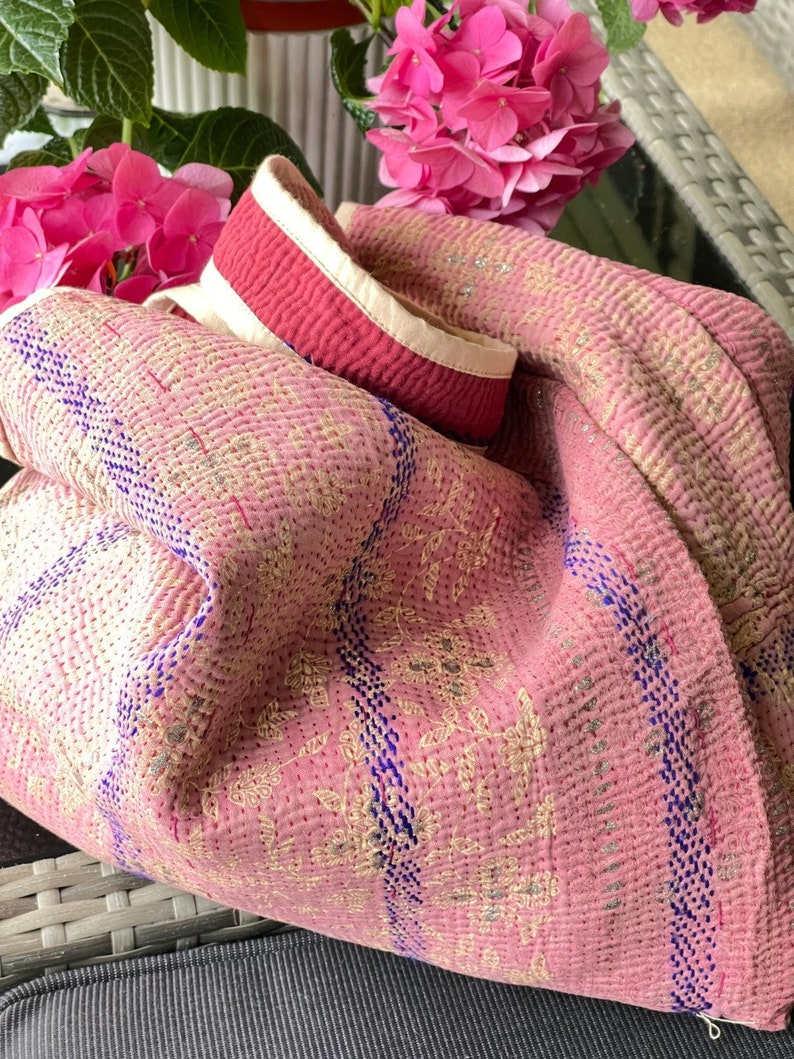 K&ouml;nnte beinhalten: Rosa gesteppte Tragetasche mit einem wei&szlig;en und roten Riemen. Die Tasche hat ein Blumenmuster und blaue N&auml;hte.