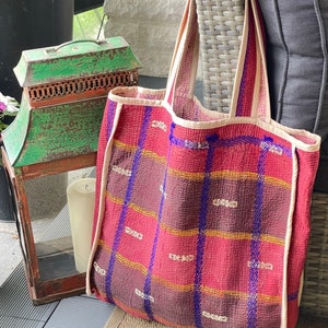 K&ouml;nnte beinhalten: Eine rote und violette karierte Tragetasche mit cremefarbenem Besatz. Die Tasche ist aus weichem, gestepptem Stoff gefertigt und hat eine gro&szlig;e, offene Oberseite.