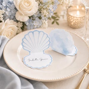 Puede incluir: Una mesa con un plato blanco, cubiertos dorados y una tarjeta de mesa en forma de concha azul. La tarjeta de mesa tiene el nombre "Isabella Grace" impreso. Un ramo de rosas blancas y hortensias azules en el fondo.