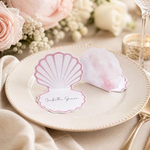 May include: A seashell-shaped place card with the name "Isabella Grace" printed on it, resting on a cream-colored plate. The card has a pink outline and a watercolor-style design. Soft pink flowers and a gold fork are in the background.