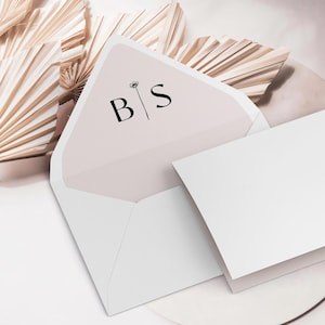 May include: A white envelope with a pink inner flap. The flap has a black floral design and the initials "B|S" printed on it. The envelope is lying on a white surface with a beige fan behind it.