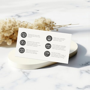 May include: A white card with black text and circular icons detailing cupcake care instructions. The card is on a white, circular pedestal, with dried flowers in the background. The card's instructions include how to transport and store cupcakes.