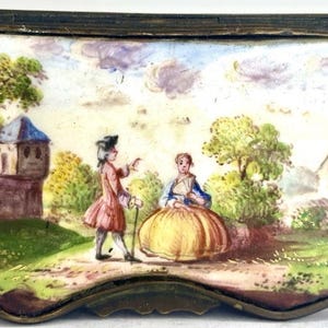 May include: Antique rectangular trinket box with a painted landscape scene. The lid features a detailed depiction of two figures in period clothing, a building, and trees. The box has a dark metal frame and a curved base.