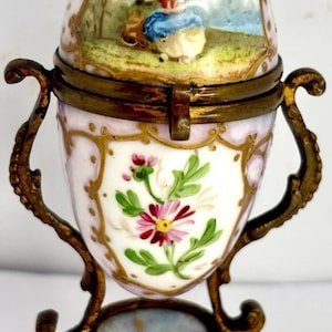 May include: An antique egg-shaped trinket box with a hinged lid, featuring a hand-painted scene of a woman in a landscape. The box is white with pink accents, gold trim, and a floral design. It rests on a decorative brass stand.