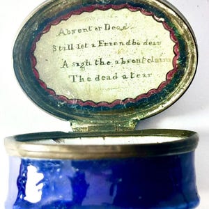 May include: An antique, oval-shaped trinket box with a hinged lid. The box is cobalt blue with a gold rim. The lid is open, revealing a handwritten poem: "Absent or Dead, Still let a Friend the dear, A sigh the absent claim, The dead a tear."
