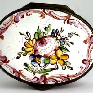 May include: Oval trinket box with a floral design. The lid features a white background with a central bouquet of pink, yellow, and blue flowers. The box has a dark metal frame and a decorative border in shades of pink and brown.