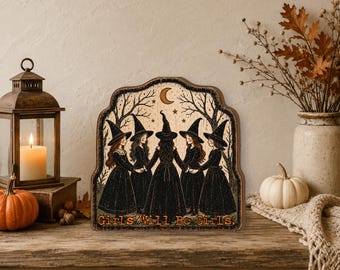 May include: A decorative wooden sign with a scalloped top edge featuring an illustration of five figures in black dresses and pointed hats holding hands in a circle under a crescent moon and stars, with the text Girls Will Be Girls printed below.