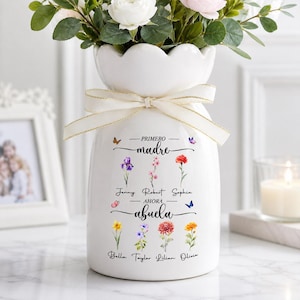 Könnte beinhalten: Weiße Keramikvase mit gewelltem Rand, gebunden mit einem cremefarbenen Band. Die Vase ist mit floralen Illustrationen und dem spanischen Text "PRIMERO madre - AHORA abuela" und Namen verziert. Die Vase enthält einen Blumenstrauß.