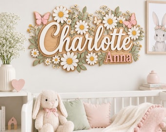 May include: A wall-mounted wooden sign featuring the names Charlotte and Anna in layered script and block lettering. The design is decorated with white daisies, green leaves, and pink butterflies, creating a floral arrangement around the text.