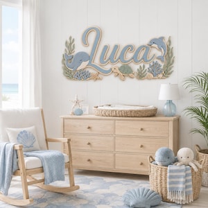 Puede incluir: Un letrero de pared de madera con el nombre Luca en letras cursivas azules, decorado con recortes en capas de una ballena, un delfín, una tortuga marina, coral y algas en tonos azul, verde y bronceado sobre una pared blanca con paneles.