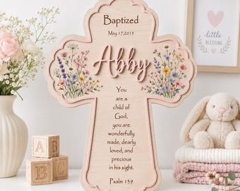 May include: A light-colored wooden cross wall plaque featuring the text "Baptized May 17, 2015", the name "Abby" in raised script, and a religious verse. The cross is decorated with colorful floral illustrations of wildflowers.