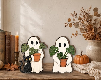 May include: Two wooden ghost figurines with white bodies and black eyes. One ghost holds a potted monstera plant and stands next to a black cat with yellow eyes. The second ghost holds a potted monstera plant. Both have dark brown edges.