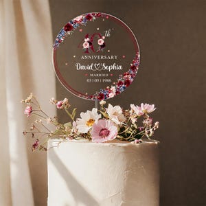 May include: A clear, circular cake topper with a floral design and the text "40th Anniversary David & Sophia Married 03 01 1986." The topper is placed on top of a white frosted cake, decorated with flowers.