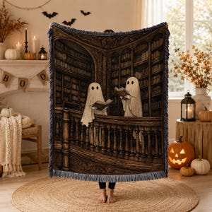 Pode incluir: Uma manta tecida com uma ilustração escura e detalhada de dois fantasmas brancos em pé numa varanda de biblioteca de madeira, cada um segurando um livro aberto. A cena inclui estantes altas e cheias ao fundo e uma borda com franjas.