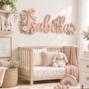 Pode incluir: Uma placa de madeira para parede exibindo o nome Isabella em letra cursiva rosa, decorada com laços rosas esculpidos, flores brancas e folhas verdes. A placa está posicionada acima de um berço de madeira clara com roupa de cama.
