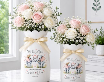 May include: Two white ceramic vases with bouquets of pink and white roses and baby's breath. Each vase has a cream-colored ribbon and a design of two elephants with the text "The Ivory Year", "14 Years Together", and names.