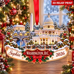 Peut inclure: Une décoration de Noël en relief 3D représentant une scène enneigée de Washington, D.C. avec le Capitole, le Washington Monument et le Lincoln Memorial. La base porte l'inscription Sophia, WASHINGTON, D.C., 2026 et un nœud rouge.