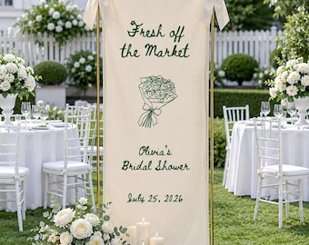 May include: A tall, rectangular cream-colored fabric banner suspended on a gold metal frame with white bows at the top corners. It features dark green embroidered text reading Fresh off the Market, Olivia's Bridal Shower, and July 25, 2026.