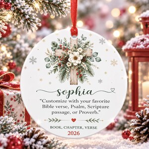 Könnte beinhalten: Ein runder, weißer Keramik-Weihnachtsschmuck mit einer Holzkreuz-Grafik, verziert mit Tannenzweigen, weißen Blüten und roten Beeren. Er enthält den Namen Sophia, den Text "Customize with your favorite Bible verse, Psalm, Scripture passage, or Proverb." sowie die Jahreszahl 2026.