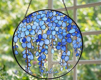 May include: Round stained glass window art featuring blue wisteria flowers. The flowers cascade downwards, with a black border and chain for hanging. The name "Sophia" is written in cursive script.
