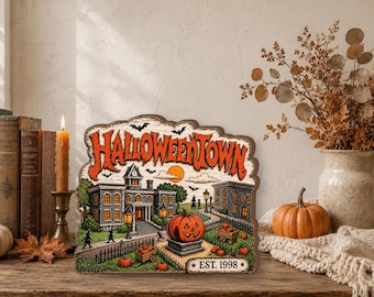 May include: A decorative wooden sign featuring a colorful illustration of a town with a large orange jack-o'-lantern in the foreground. The text HALLOWEENTOWN is printed in large orange letters at the top, with EST. 1998 at the bottom right.