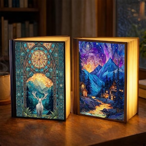 May include: Two open book-shaped lamps with illuminated pages. One features a stained-glass design with a deer, the other a mountain landscape. The lamps have black covers and are set on a wooden surface, creating a warm, inviting ambiance.