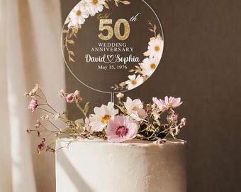 May include: A clear acrylic cake topper for a 50th wedding anniversary. The topper is round with gold text that reads "50th Wedding Anniversary David & Sophia May 15, 1976". It is decorated with white and gold floral accents.