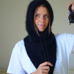 May include: A black, fluffy hood with a drawstring closure. The hood is worn over a white t-shirt. The hood appears to be made of a soft, textured material. The overall style is casual and warm.