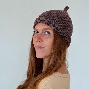 May include: A brown knit beanie with a small, rounded top detail. The hat is made of a ribbed knit fabric and is worn on the head. The person in the image is wearing a tan colored shirt.
