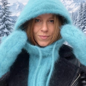 May include: A light blue, fluffy hood and matching mittens are worn over a black jacket. The hood is part of a scarf that wraps around the neck. The background shows a snowy mountain scene.