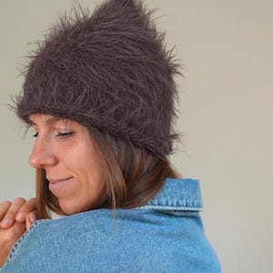 May include: A dark brown beanie with a fuzzy, textured surface and a pointed top. The hat features a knitted construction with long, soft fibers that create a shaggy appearance. The beanie is worn low on the forehead, covering the ears completely.