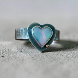 May include: A silver ring featuring a heart-shaped opal stone. The opal displays a mix of blue, green, and pink hues. The ring has a textured band and a small, gold-colored accent near the heart.