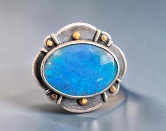 May include: An oval ring featuring a vibrant blue stone set in a textured silver frame. The silver setting is accented with small, raised gold-toned circular studs positioned around the perimeter of the central blue oval gemstone.