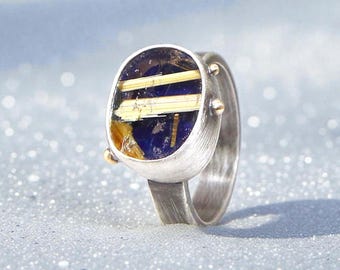 May include: A silver ring with an oval, dark blue stone inlaid with gold-colored bars. The ring has a brushed metal finish and small gold accents on the sides of the stone setting. The ring is set against a light, textured background.