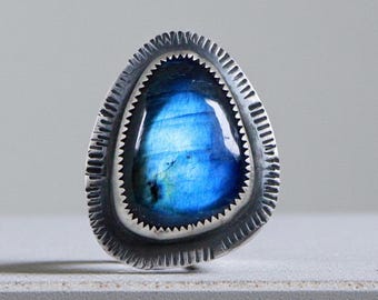 May include: A ring featuring a triangular labradorite gemstone with a vibrant blue flash, set in a silver bezel. The metal surrounding the stone has a textured, serrated edge with vertical etched lines, creating a distinct border around the gem.