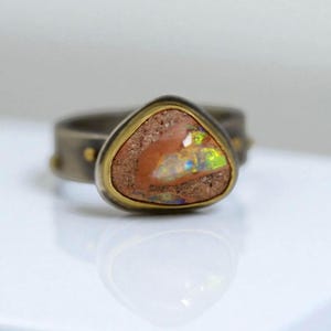 May include: A close-up of a ring featuring a triangular, earthy-toned opal stone with iridescent flashes of color. The stone is set in a gold bezel within a dark metal band with small gold accents. The ring is on a white surface.