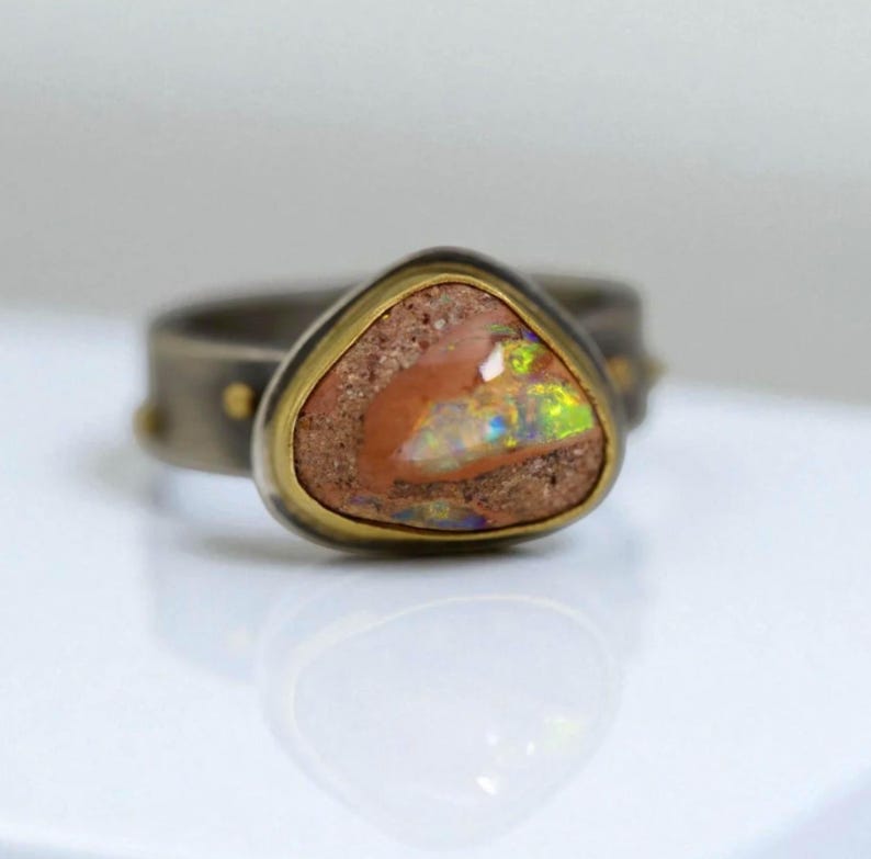 May include: A close-up of a ring featuring a triangular, earthy-toned opal stone with iridescent flashes of color. The stone is set in a gold bezel within a dark metal band with small gold accents. The ring is on a white surface.