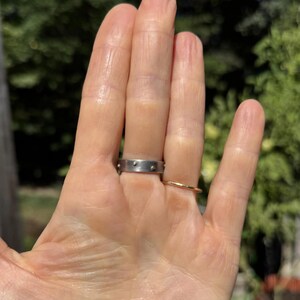May include: A hand wearing two rings. One is a silver band with small gold-colored studs. The other is a thin, gold-colored band. The background is a blurred view of green foliage.