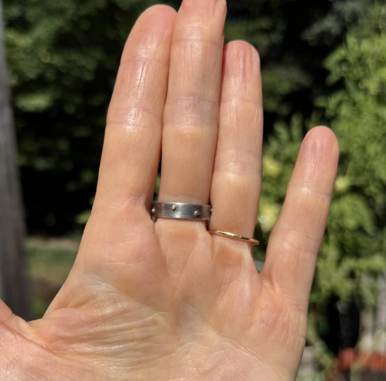 May include: A hand wearing two rings. One is a silver band with small gold-colored studs. The other is a thin, gold-colored band. The background is a blurred view of green foliage.