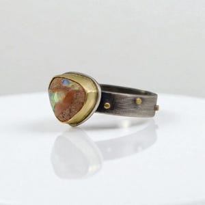 May include: A handcrafted ring featuring a triangular, multi-colored opal stone set in a gold bezel. The band is made of oxidized silver with small gold accents. The ring is a unique piece of jewelry.