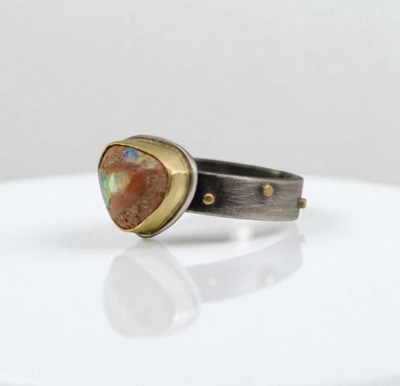 May include: A handcrafted ring featuring a triangular, multi-colored opal stone set in a gold bezel. The band is made of oxidized silver with small gold accents. The ring is a unique piece of jewelry.