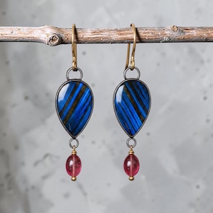 May include: A pair of drop earrings featuring teardrop-shaped blue and black striped stones set in dark metal frames. Each earring hangs from a gold-toned hook and includes a small, round, translucent pink bead suspended at the bottom.
