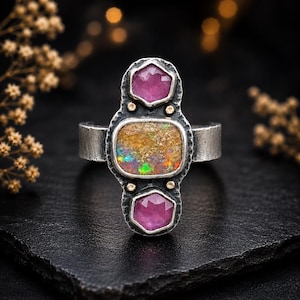 May include: A silver ring featuring a central rectangular opal with iridescent colors, flanked by two pink, hexagonal gemstones. The ring has a textured band and small gold accents. The ring is displayed on a black surface.