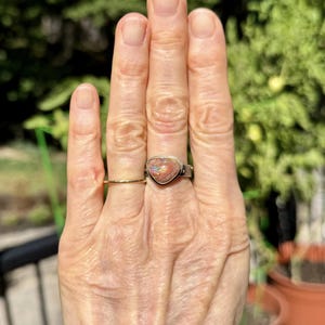 May include: A silver ring with a triangular, multi-colored stone and a gold band ring. The stone has shades of red, orange, and blue. The rings are on a hand with a blurred green background.