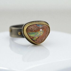 May include: A unique ring featuring a triangular, multi-colored opal stone set in a gold bezel. The band is a dark, textured metal with small gold accents. The opal displays shades of brown, green, and blue.