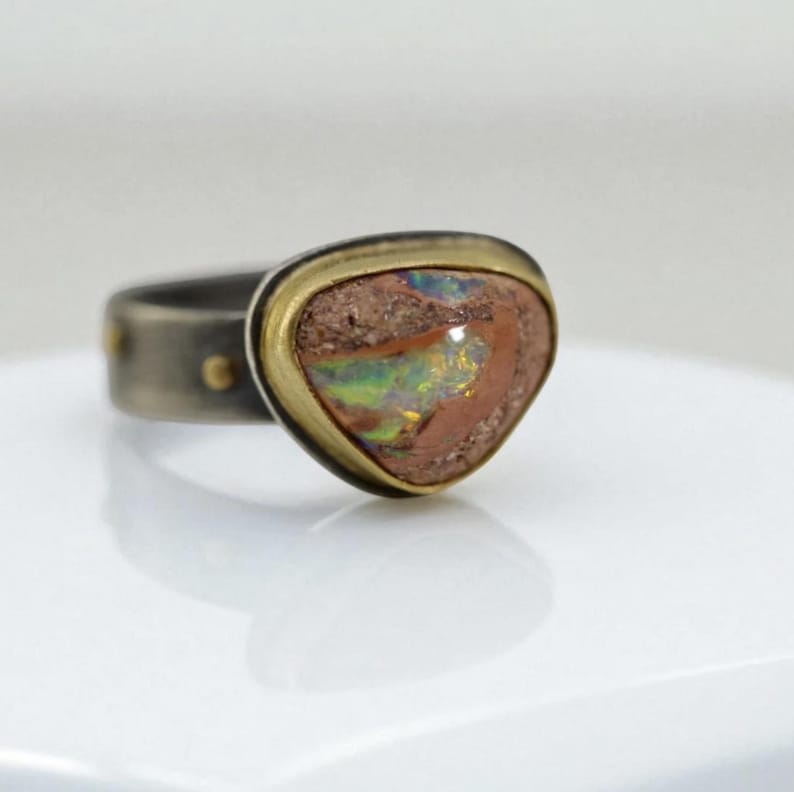 May include: A unique ring featuring a triangular, multi-colored opal stone set in a gold bezel. The band is a dark, textured metal with small gold accents. The opal displays shades of brown, green, and blue.