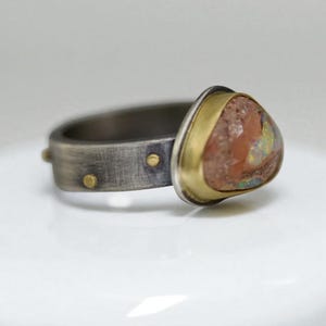 May include: A handcrafted ring featuring a triangular, multi-colored opal stone set in a gold bezel. The band is made of silver-toned metal with a textured finish and small gold accents. The ring is a unique piece of jewelry.