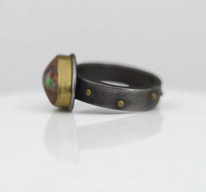 May include: A close-up of a ring with a large, oval-shaped stone. The stone has a mix of colors, including red, green, and brown. The ring's band is dark gray with small gold accents. The setting around the stone is gold.